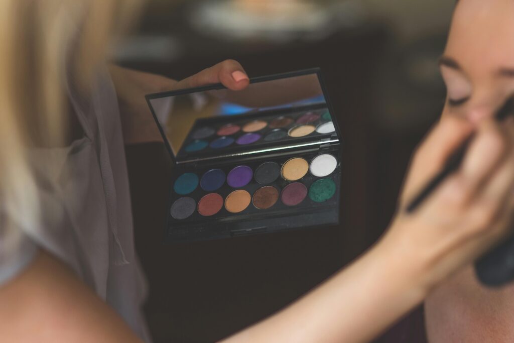 A close-up of a stylist applying eyeshadow from a vibrant color palette to a model's face indoors.